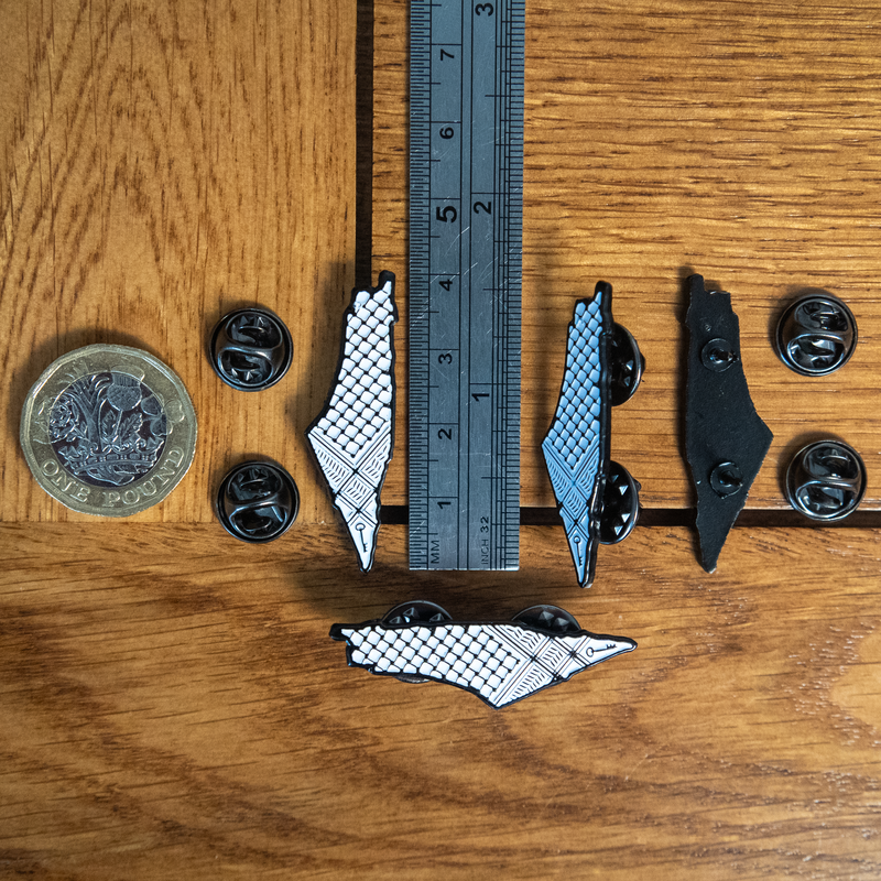 Image of the Keffiyeh Enamel Pin aligned next to a ruler showing its 40mm length, as well as a pound coin for scale. Other pins are also in the picture including the back of the pin showing the double-pins included for stability.